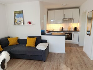 A modern living room with a grey sofa and yellow cushions, a small white table, and a white fluffy chair. A kitchen with white cabinets and a wooden countertop is visible in the background. - Modern Vacation Apartment Silvia Near Lake Constance (Weissensberg / Lindau (Bodensee))