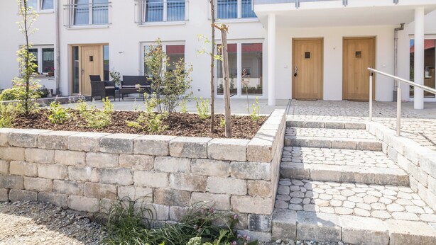 Un edificio de varios pisos con un muro de contención de piedra, asientos al aire libre y una escalera que lleva a la entrada.