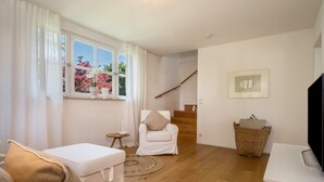 A living room with a white sofa, a white armchair, a wooden floor, a staircase, a wicker basket, and a framed picture on the wall. - "Holiday cottage Fischbach" close to the Lake with Wi-Fi (Friedrichshafen)