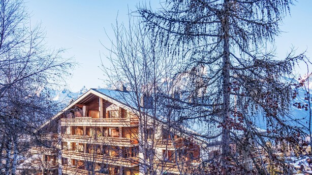 En stor, flervånings chalet byggd av trä, med balkonger, omgiven av träd och med berg i bakgrunden.