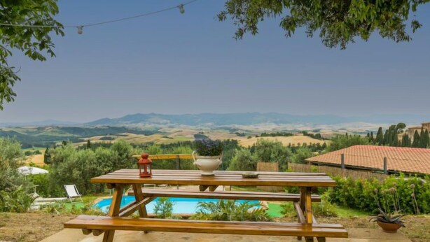 A wooden picnic table with a white pot and a red lantern on it, overlooking a swimming pool and a scenic landscape with hills and trees. - Miranda by Interhome (Volterra)