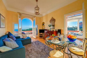 Amplia sala de estar con un sofá azul, mesa de comedor y sillas, zona de cocina y ventanas grandes con vistas al exterior.