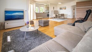 Un salon moderne avec un grand canapé, une table basse et un tableau au mur. On y trouve également un coin repas avec une table et des chaises, et une cuisine à l’arrière-plan.