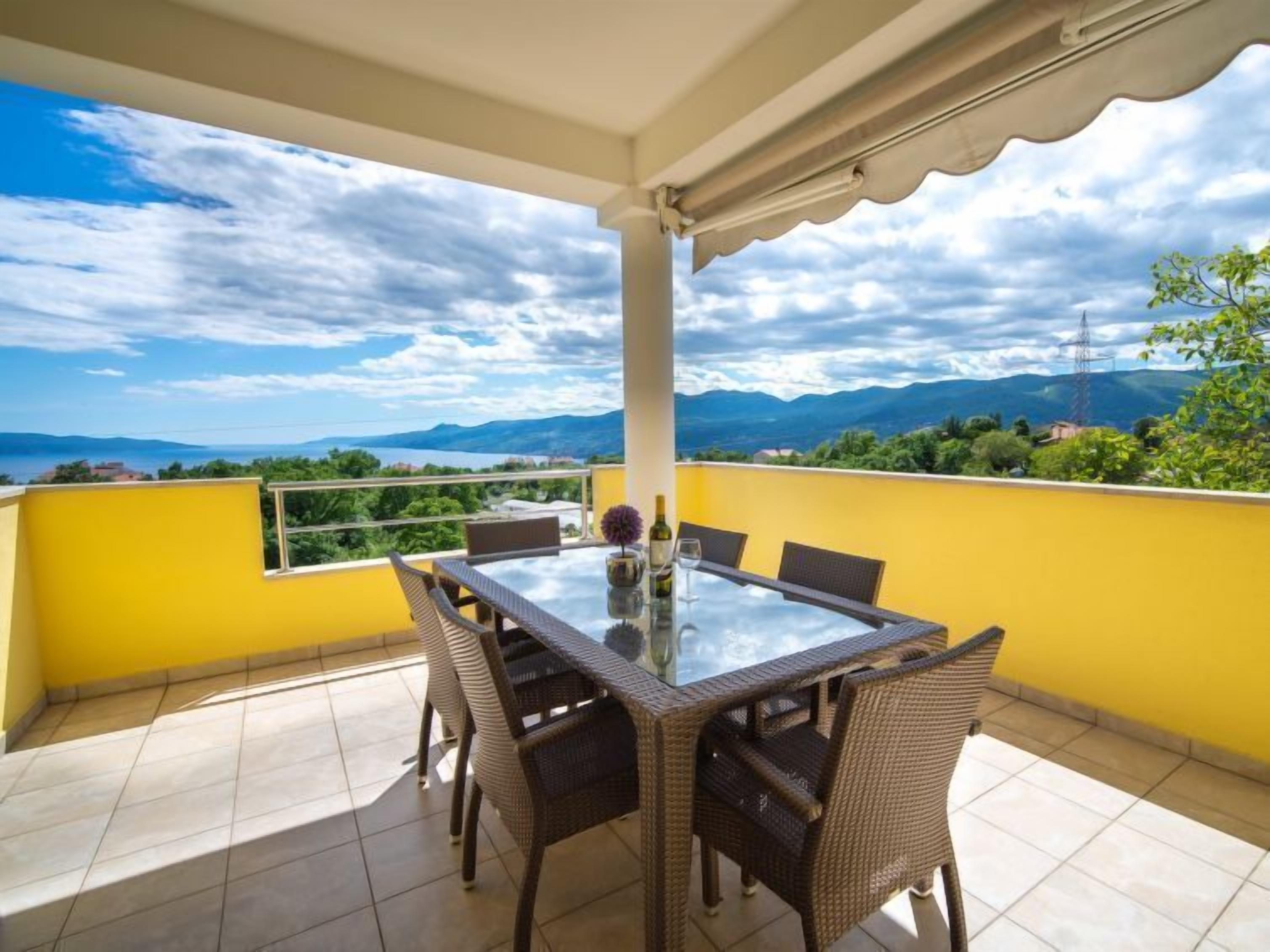 A balcony with a table and chairs, a glass-top table, and a view of mountains and greenery.