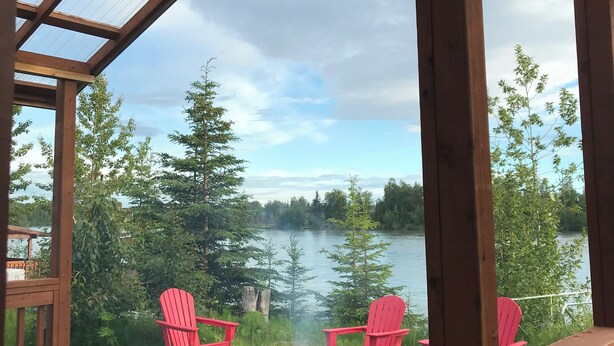 Une terrasse en bois avec un toit de verre donnant sur un lac, deux chaises Adirondack rouges et un barbecue.