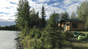 Une cabane au bord d’une rivière, avec des kayaks et une forêt dense à proximité.