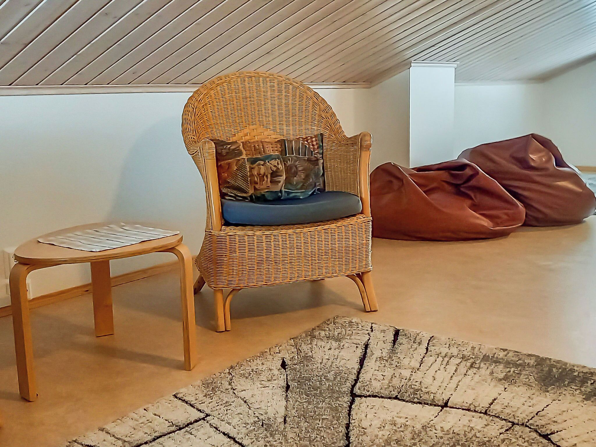 A wicker chair with a cushion, a small round table, and a bean bag chair in a room with a sloped ceiling and a patterned rug.