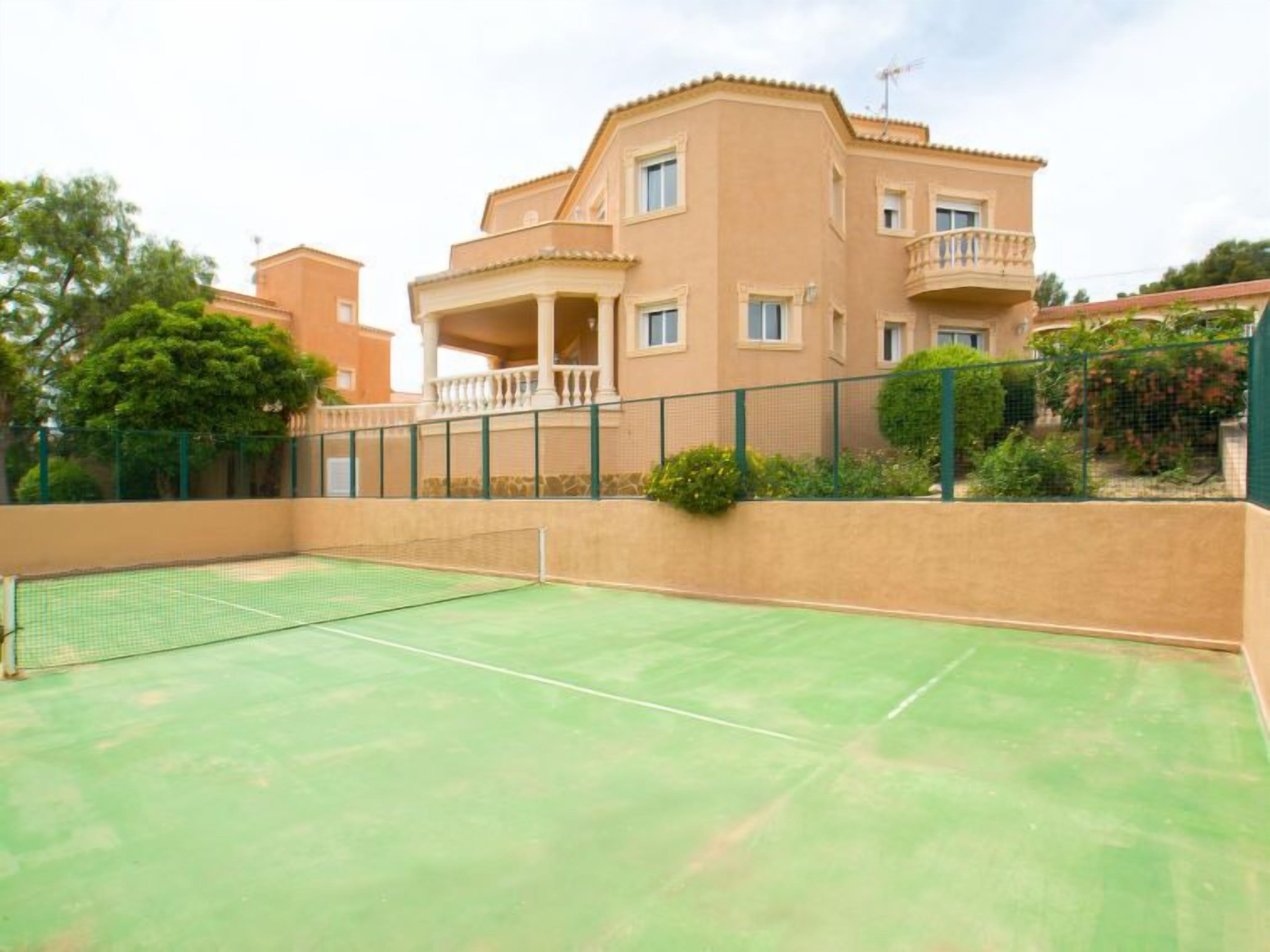 Un court de tennis extérieur, situé à côté d’un immeuble à plusieurs étages doté de balcons et entouré d’une clôture verte.