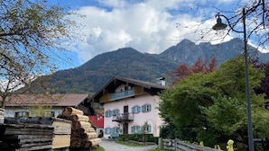 A rural mountain road leading to a traditional chalet with a balcony, surrounded by greenery and mountains.