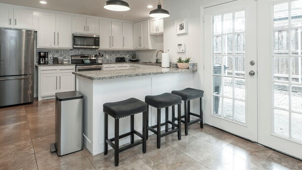 Una cocina moderna con armarios blancos, electrodomésticos de acero inoxidable, una isla central con taburetes y un amplio ventanal con vistas al exterior.