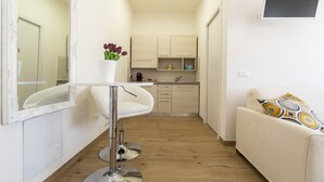 A narrow, well-lit corridor with a white sofa and a small table with a vase of flowers. A kitchenette with cabinets and a sink is visible in the background. - Liberty Fiera Bologna (Bologna)