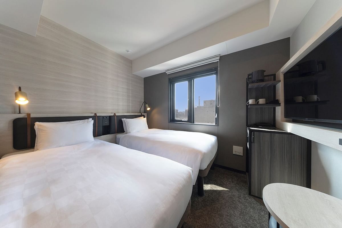Ein Hotelzimmer mit zwei Betten, einem Schreibtisch, einem Fernseher und einem Fenster mit Blick auf die Stadt.