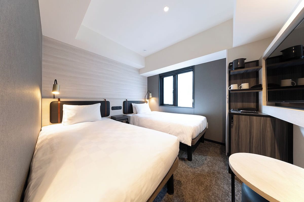 Ein Hotelzimmer mit zwei Betten, einem kleinen Tisch, einem Regal mit Tassen und einem Fenster.