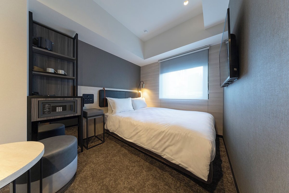 Ein Hotelzimmer mit einem Bett, einem Schreibtisch mit Stuhl, einem kleinen Tisch, einem Bücherregal und einem an der Wand montierten Fernseher.
