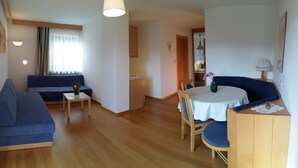 En stue med sofa, spisebord med stole, et lille tekøkken og et vindue med gardiner.
