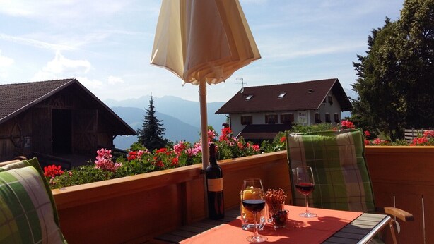 En terrasse med et spisebord, vin og glas, omgivet af blomster og udsigt til et bjerg.