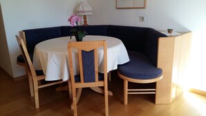 Et spisebord i hjørnet med runde bordplade, to stole, en vase med blomster, en lampe og et indrammet billede på væggen.