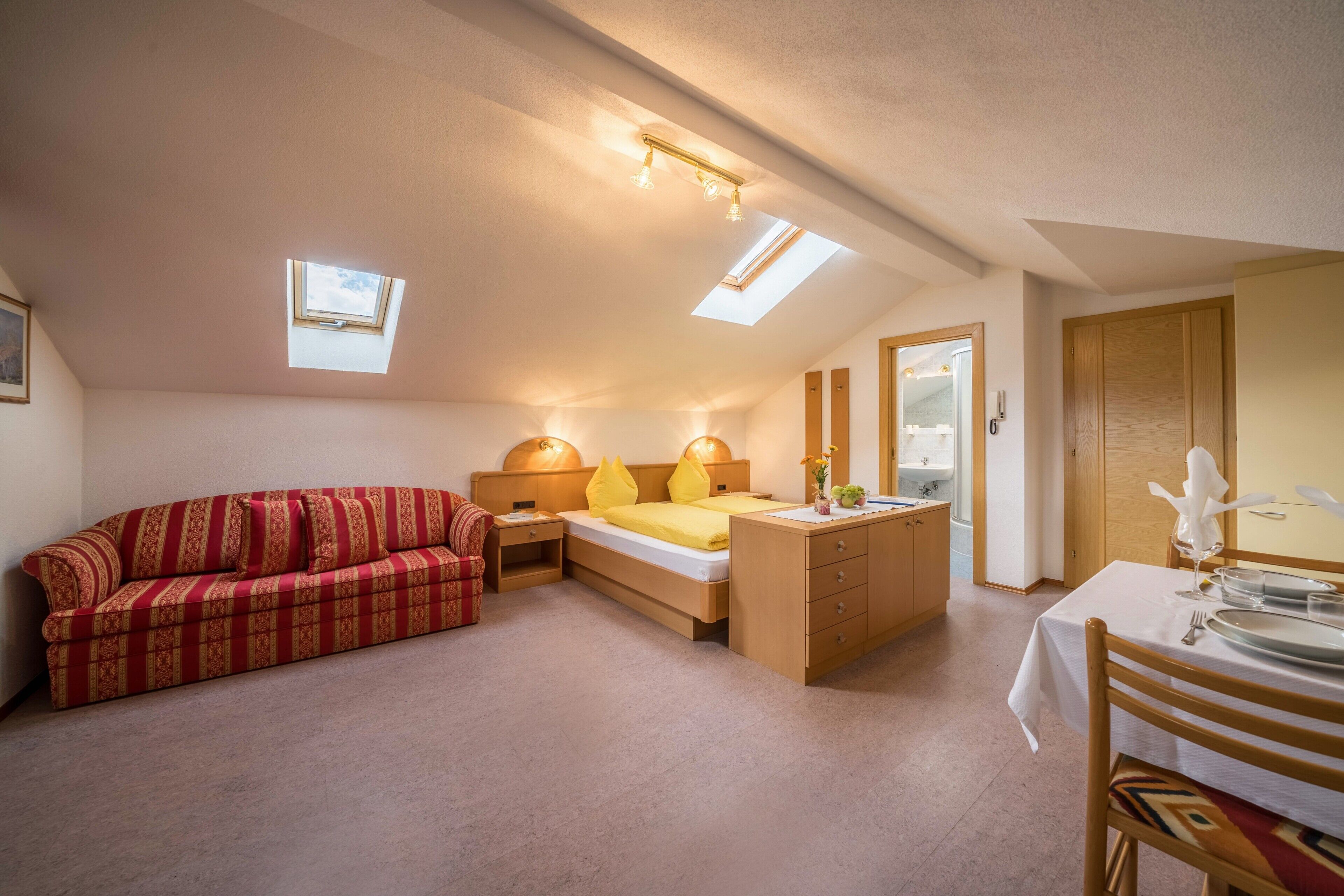 Une chambre à coucher comprenant un lit, un canapé, un bureau avec une chaise et une petite table avec une lampe. Il y a des fenêtres de toit et une porte qui donne sur une autre pièce.