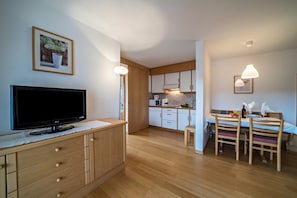 A modern kitchen with wooden cabinets, a dining area with a table and chairs, a TV on a cabinet, and pendant lighting.