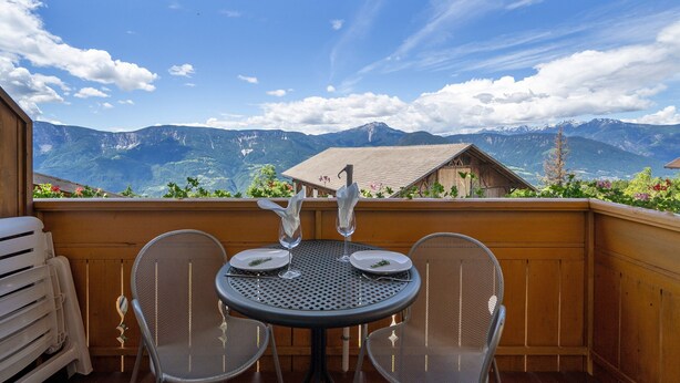 Un balcone in legno con un tavolo rotondo e due sedie, con vista su un paesaggio montuoso.