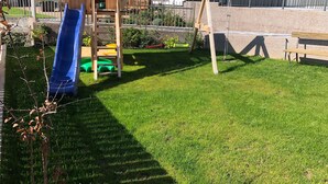 Un cortile con una struttura giochi in legno, uno scivolo blu e un’altalena.