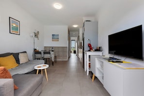 En moderne stue med en grå sofa, et lille rundt bord, et fjernsyn på et stativ og et køkken i baggrunden.