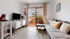 Ein modernes Wohnzimmer mit einem weißen Sofa, einem Fernseher, einem kleinen Tisch und einer Glastür, die auf einen Balkon im Freien führt.