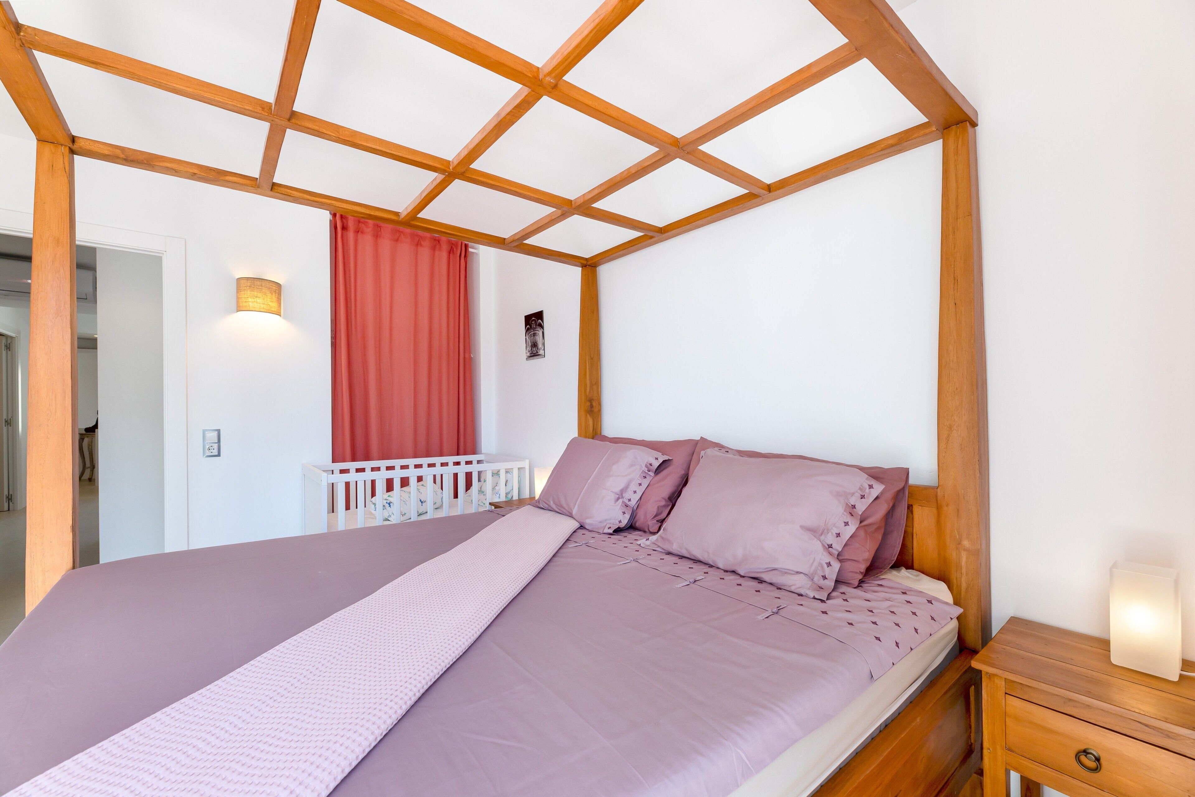 Un dormitorio con techo de madera, una cama con colcha rosa, una mesita de noche con lámpara y una cortina roja.