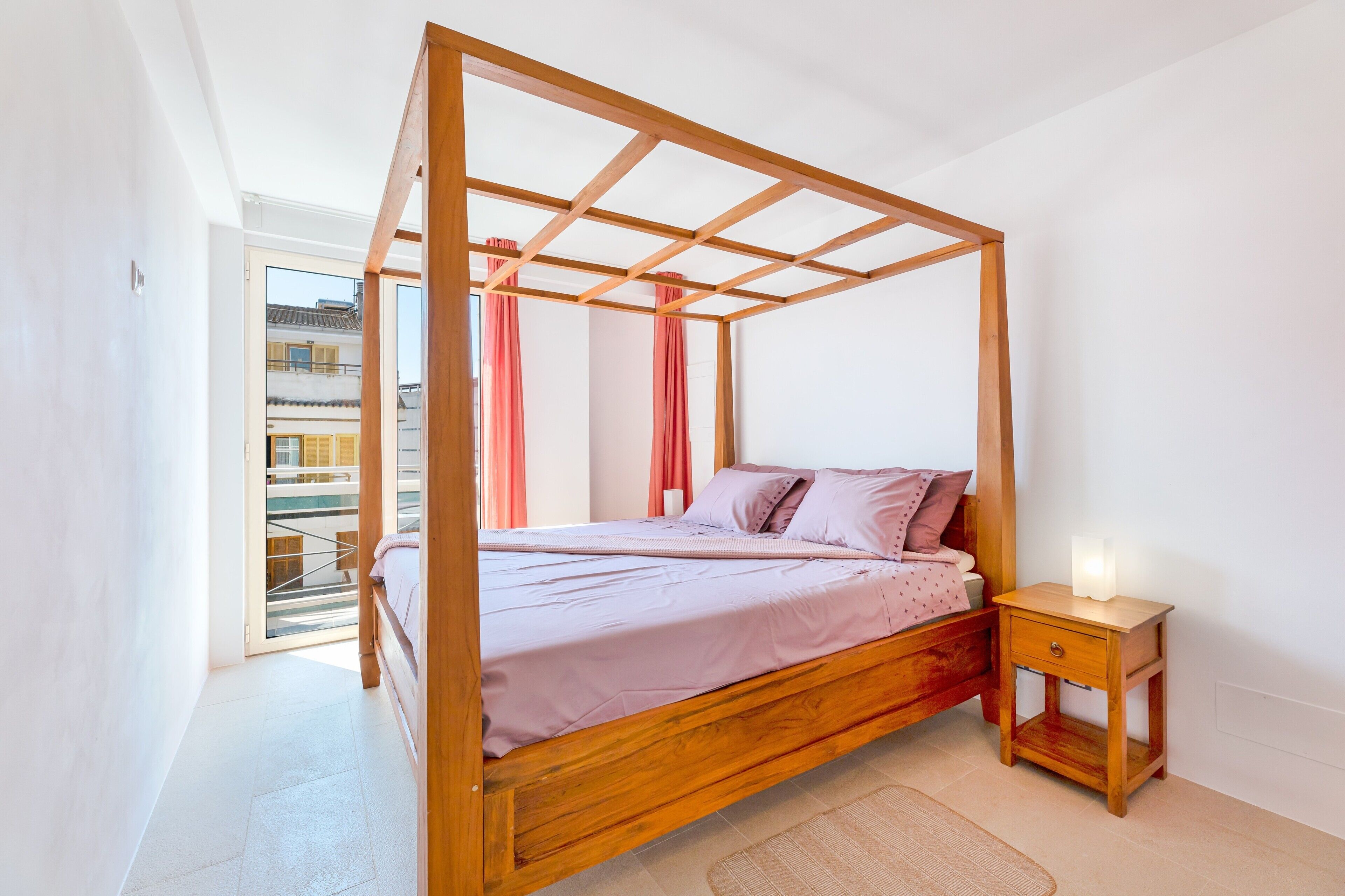 A bedroom with a four-poster bed, a bedside table with a lamp, and a view of a building through a glass door.