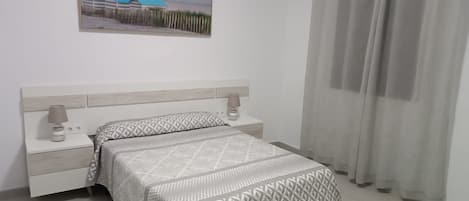Quarto com cama, mesas de cabeceira, luminária, quadro e janela com cortinas.
