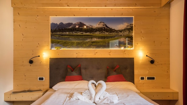 Une chambre à coucher avec un lit, une décoration en forme de cœur réalisée avec des serviettes, un tableau représentant un paysage de montagne au-dessus du lit, et deux tables de chevet avec des lampes.
