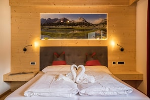 Une chambre à coucher avec un lit, une décoration en forme de cœur réalisée avec des serviettes, un tableau représentant un paysage de montagne au-dessus du lit, et deux tables de chevet avec des lampes.