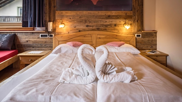 A hotel room with a bed featuring two towels arranged in swan shapes, a wooden headboard, a painting of a sunset, and a window with curtains. - Pet-friendly Apartment "Villa Anita Pares" with Mountain View, Sauna, Wi-Fi, Terrace & Balcony (St.Vigil)