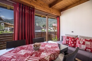 Ein Zimmer mit Holzboden, einem Tisch mit roter Tischdecke, einer Couch mit Kissen in Herzmuster und Blick auf Berge und Häuser durch große Fenster.