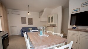 A compact living space with a dining table, white chairs, a blue sofa, a kitchen area with cabinets, and a TV mounted on the wall. - Villetta Clara, holiday house close to the beach (Nardo)