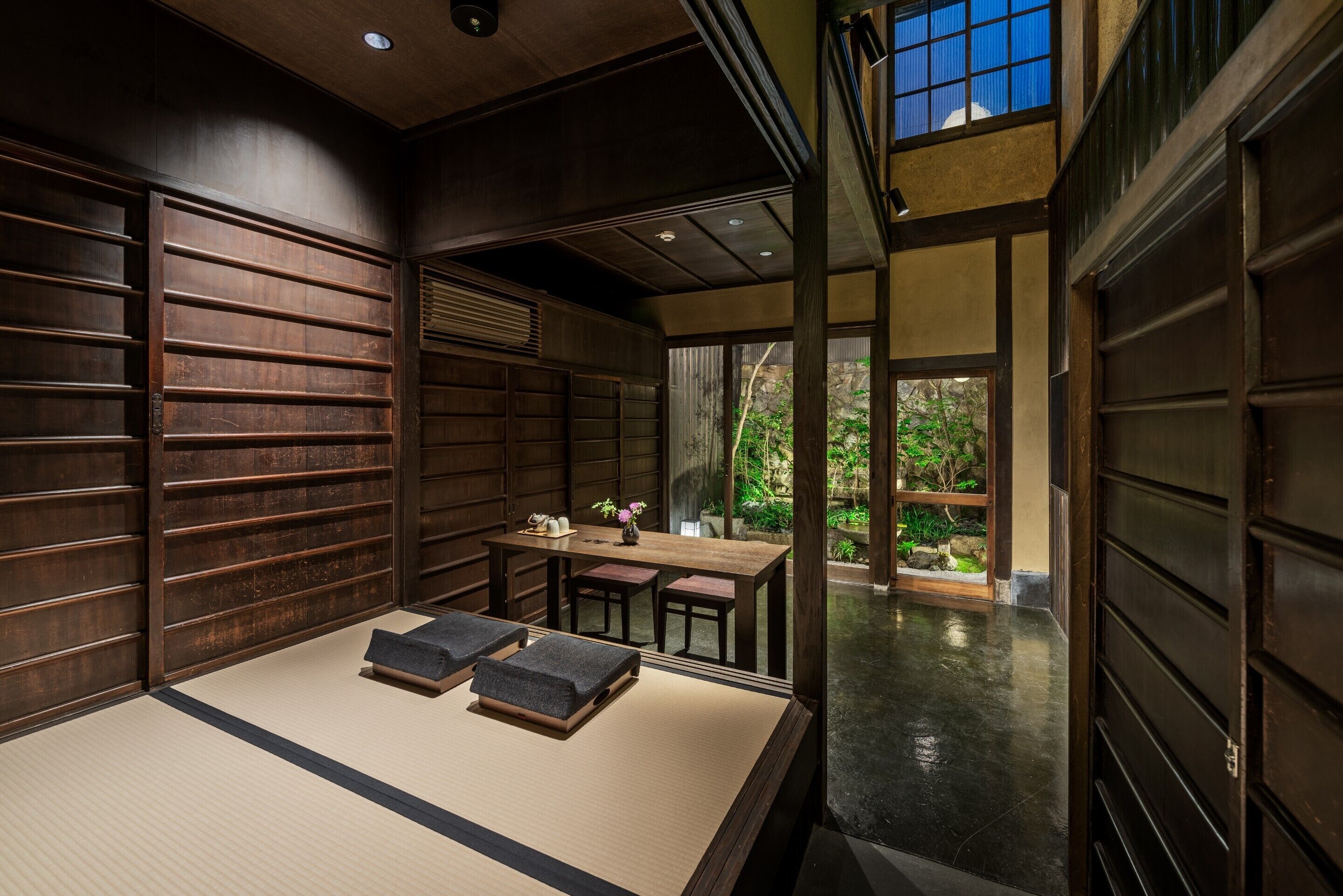 Une pièce de style japonais traditionnel, avec un sol recouvert de tatamis, une petite table basse avec des coussins et une vue sur la verdure à l’extérieur.