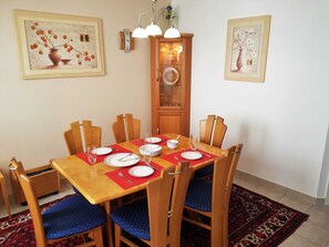 A dining area with a wooden table set for four, surrounded by chairs with blue cushions. There are framed artworks on the walls, a wooden cabinet with glass doors, and a patterned rug on the floor.
