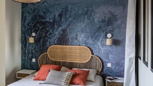Un dormitorio con una cama grande, cabecera trenzada, pared azul con textura, dos mesitas de noche, lámpara colgante y una ventana con cortinas.