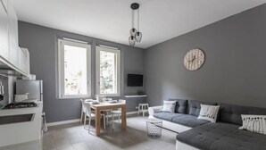 A modern kitchen with white cabinetry, a dining area with a wooden table and chairs, a gray sofa, and a large window providing natural light.