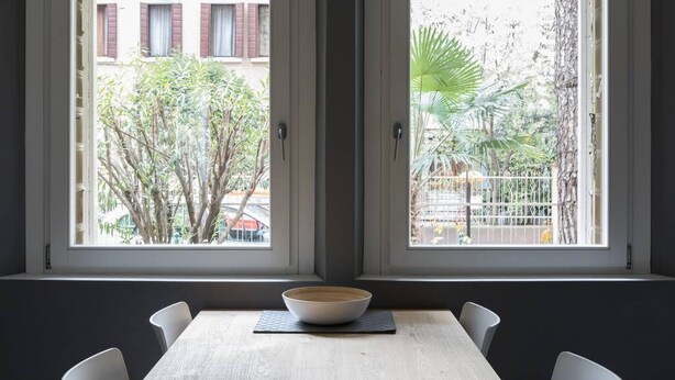 A dining area with a table and chairs, a bowl on the table, and a view of a tree and building outside through large windows.