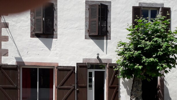 Un edificio con dos puertas de madera y ventanas con contraventanas, un árbol en primer plano y un muro de piedra.
