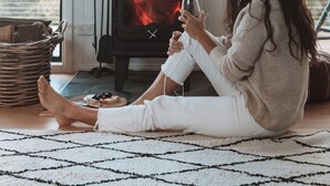 Une personne est assise sur le sol devant un poêle à bois. On aperçoit en arrière-plan une chaise en osier et une fenêtre.