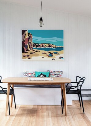 A dining area with a wooden table, black chairs, a colorful painting of a beach scene, and a hanging light fixture. - A cosy getaway you won't want to leave (Police Point)