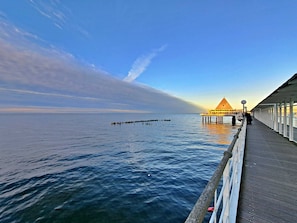 Eine Steg, der sich über ruhiges Wasser erstreckt, mit einem Gebäude am Ende, das ein spitzes Dach und eine Reihe Fenster an der Seite hat.