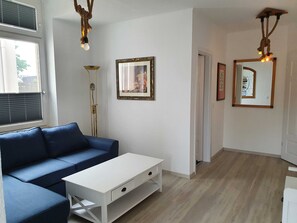 A modern living room with a blue sofa, a white coffee table, and a tall floor lamp. There are two framed pictures on the wall and a mirror with lights.