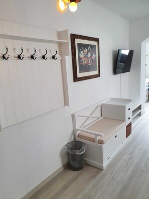 Ett rum med en vit vägg, en upphängd bildram, en tv, en bänk med lådor och en papperskorg.