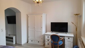 A room with a white desk, a television, a blue chair, a wardrobe, and a chandelier. - Junior Suite - Domkes Hotel Garni Haus an der See (Heringsdorf)