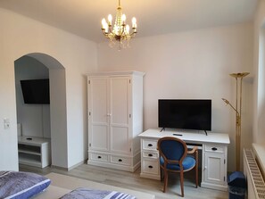 Ein Zimmer mit einem weißen Schreibtisch, einem Fernseher, einem blauen Sessel, einem Kleiderschrank und einer Deckenleuchte.