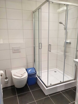 Ein modernes Badezimmer mit einer gläsernen Duschkabine, weißen Fliesen und einer weißen Toilette sowie einem blauen Mülleimer.
