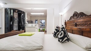 A bedroom with a bed, pillows, a wooden headboard, a bathroom with a shower, and a kitchenette in the background.